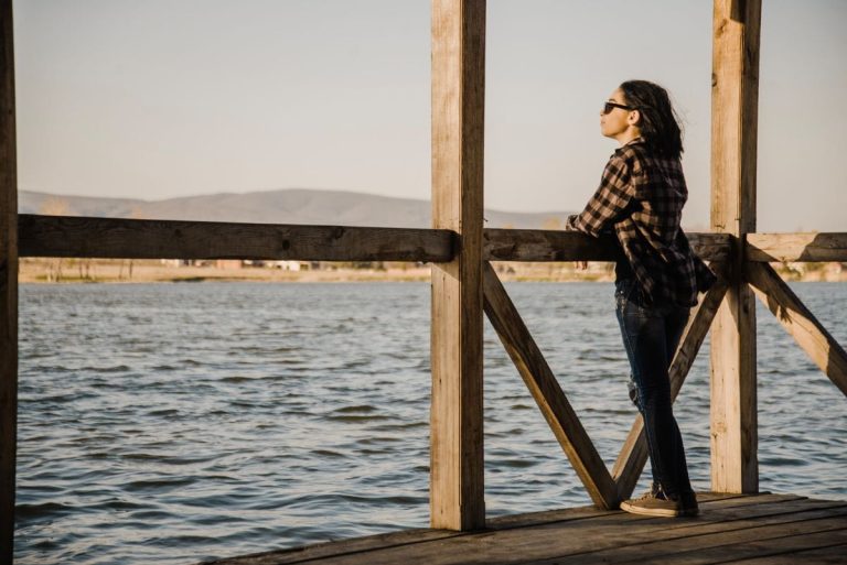 Eine Person steht allein am Meer oder auf einer Brücke und schaut ruhig in die Ferne.